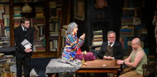 Kilka osób wokół stołu, w tle bardzo dużo książek. Scena ze spektaklu "Tango" na podstawie dramatu Sławomira Mrożka.