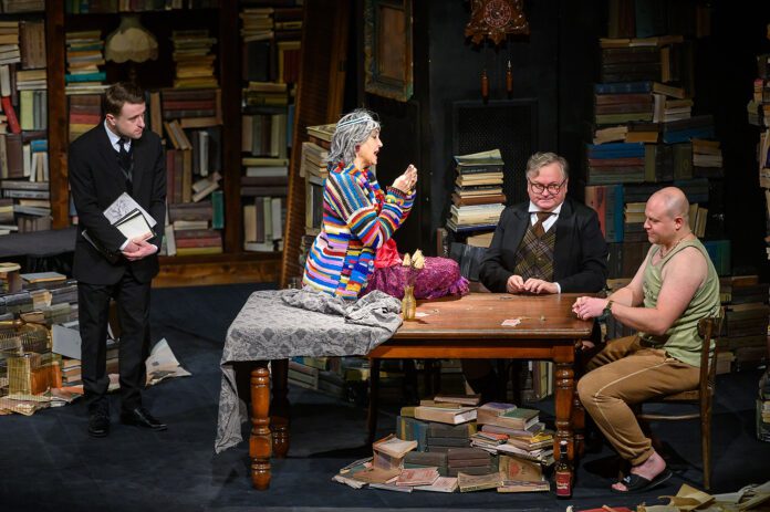 Kilka osób wokół stołu, w tle bardzo dużo książek. Scena ze spektaklu "Tango" na podstawie dramatu Sławomira Mrożka.