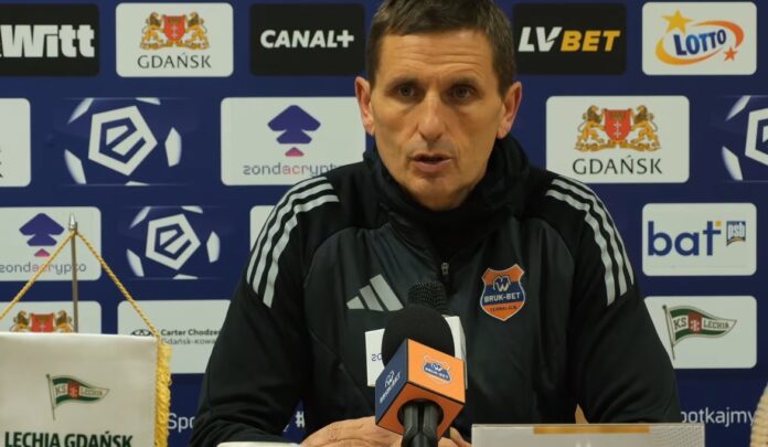 Trener Bruk-Bet Termaliki Nieciecza Marcin Brosz wypowiada się do mikrofonu na tle ścianki reklamowej podczas konferencji prasowej po meczu PKO BP Ekstraklasy